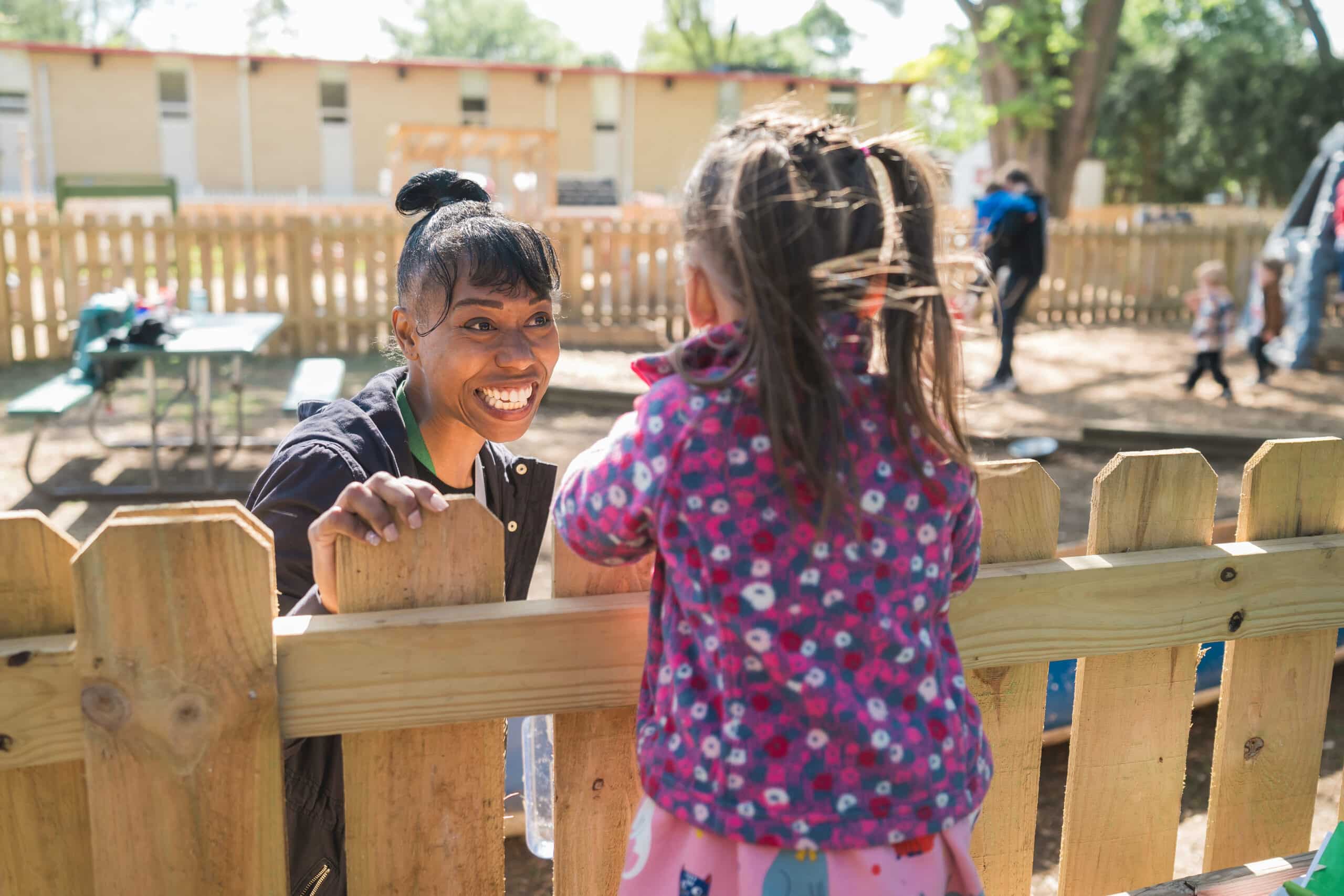 Teacher smiling and speaking with a child across an outdoor fence