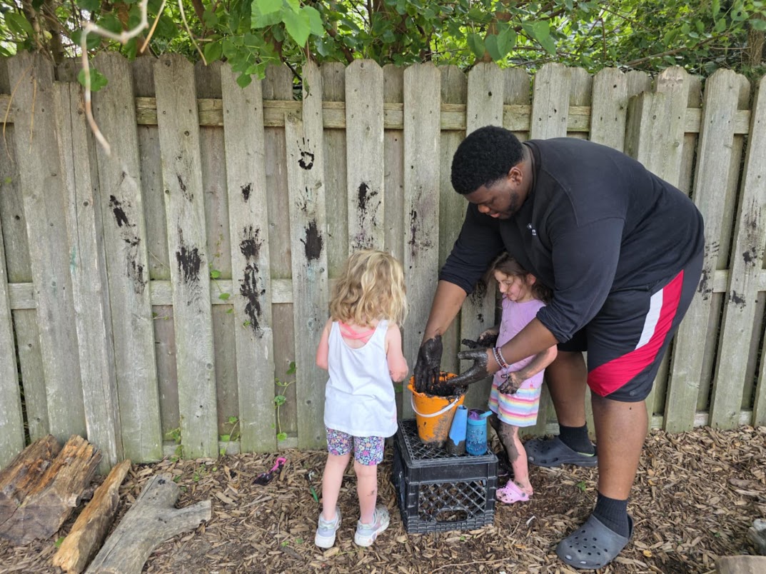 An educator and two children working with mud and natural materials outdoors
