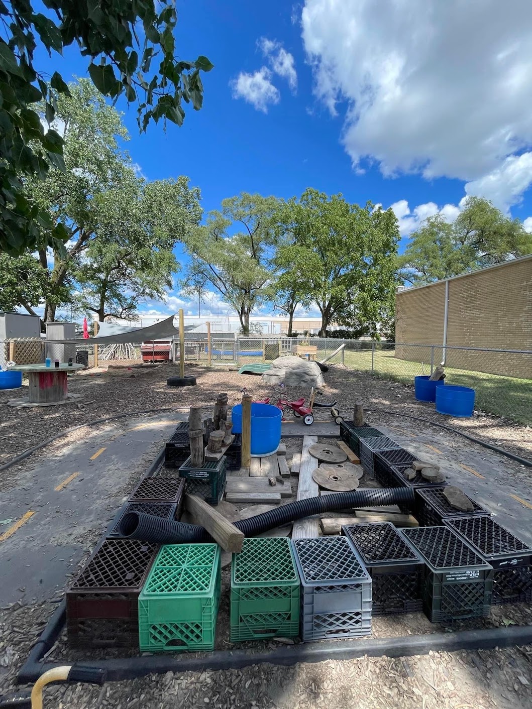 Outdoor play space arranged with milk crates, wooden pieces, round wood slices, and tubing